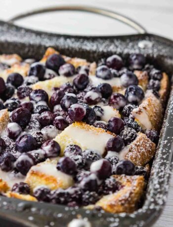 A delectable blueberry French toast casserole, soaked in a rich cream cheese egg mixture and baked to a golden brown in a metal casserole dish. The blueberries have burst with vibrant color, adding a pop of freshness to the crusty surface