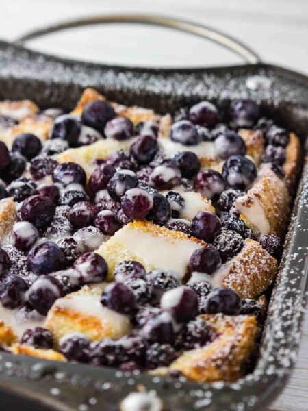 A delectable blueberry French toast casserole, soaked in a rich cream cheese egg mixture and baked to a golden brown in a metal casserole dish. The blueberries have burst with vibrant color, adding a pop of freshness to the crusty surface