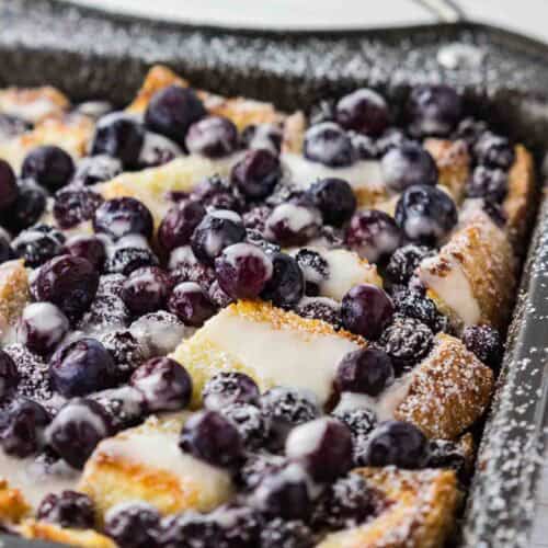 A delectable blueberry French toast casserole, soaked in a rich cream cheese egg mixture and baked to a golden brown in a metal casserole dish. The blueberries have burst with vibrant color, adding a pop of freshness to the crusty surface