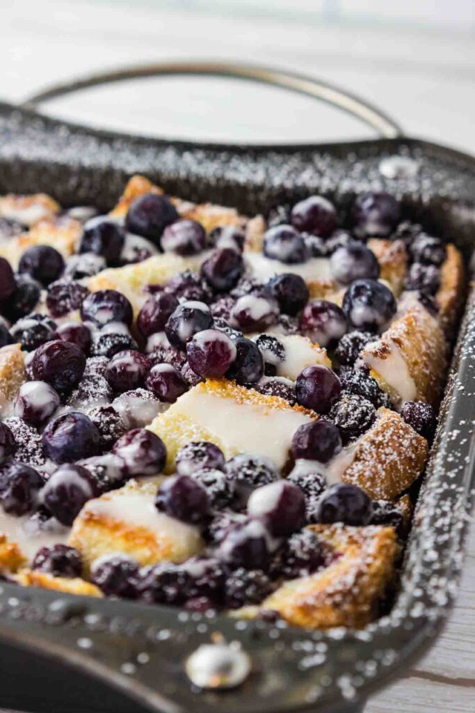 A delectable blueberry French toast casserole, soaked in a rich cream cheese egg mixture and baked to a golden brown in a metal casserole dish. The blueberries have burst with vibrant color, adding a pop of freshness to the crusty surface