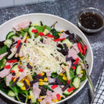 mixed green salad with corn, cucumbers, shaved Swiss cheese, ham and tricolored tortilla strips in a white bowl on countertop