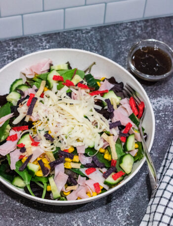 mixed green salad with corn, cucumbers, shaved Swiss cheese, ham and tricolored tortilla strips in a white bowl on countertop