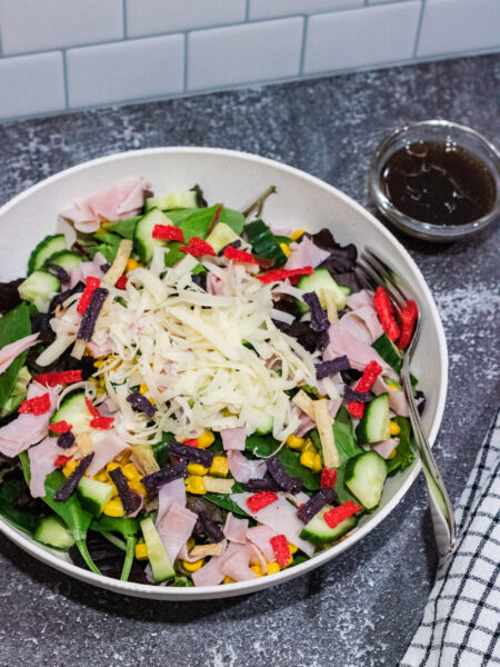 mixed green salad with corn, cucumbers, shaved Swiss cheese, ham and tricolored tortilla strips in a white bowl on countertop