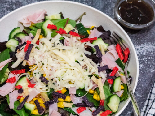 mixed green salad with corn, cucumbers, shaved Swiss cheese, ham and tricolored tortilla strips in a white bowl on countertop