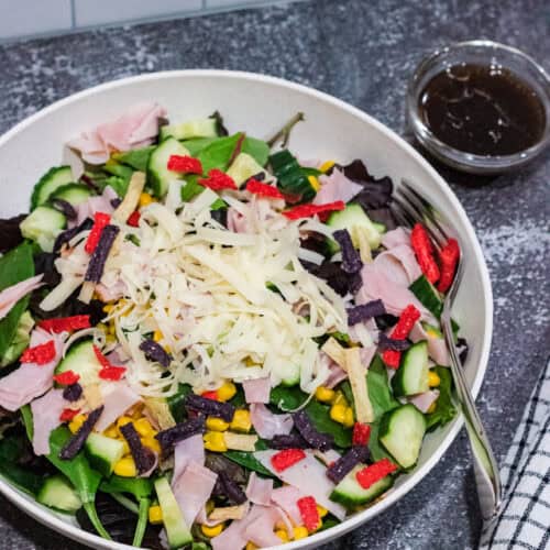 mixed green salad with corn, cucumbers, shaved Swiss cheese, ham and tricolored tortilla strips in a white bowl on countertop