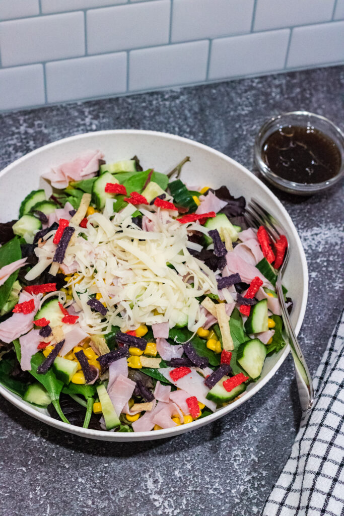 mixed green salad with corn, cucumbers, shaved Swiss cheese, ham and tricolored tortilla strips in a white bowl on countertop