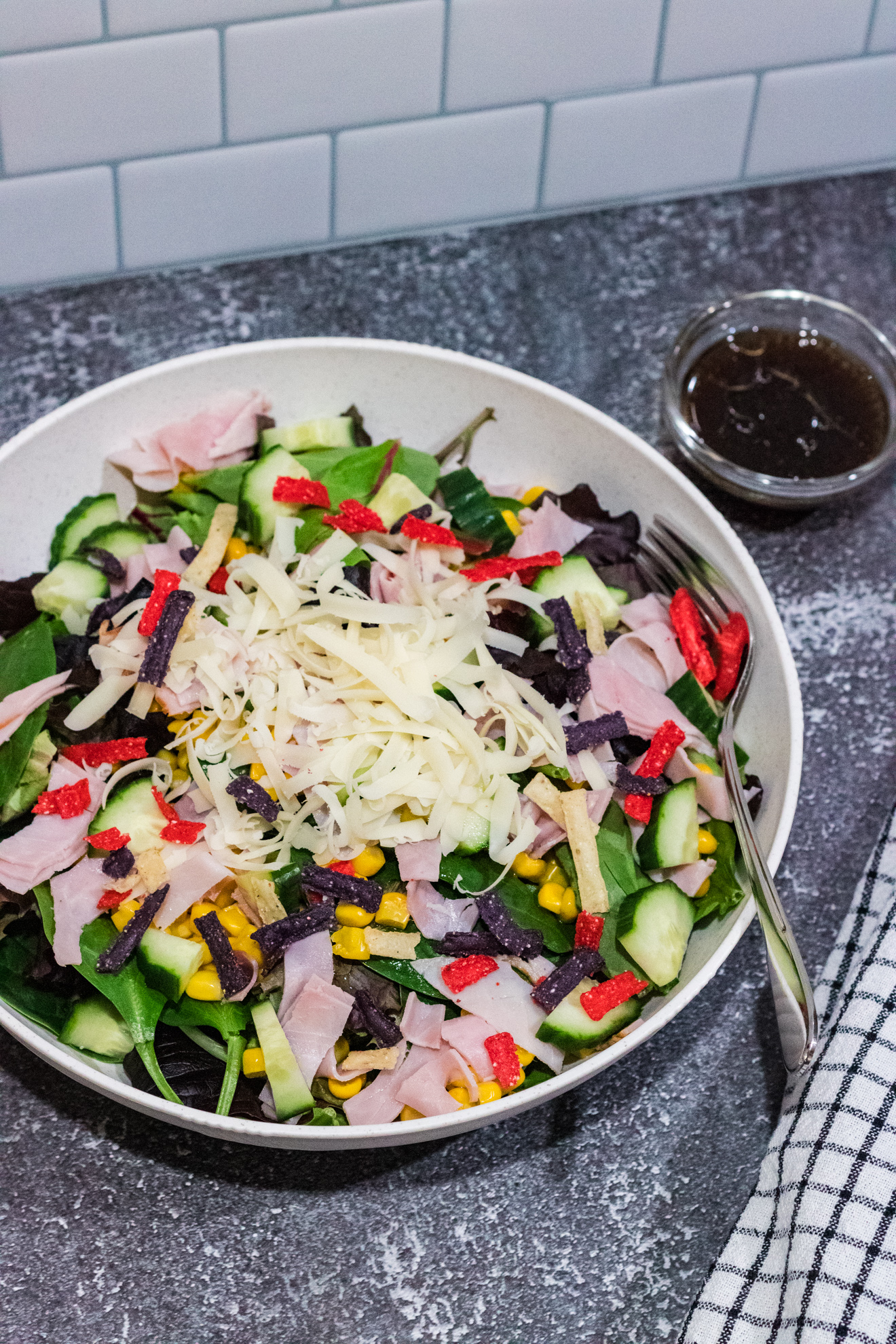 mixed green salad with corn, cucumbers, shaved Swiss cheese, ham and tricolored tortilla strips in a white bowl on countertop