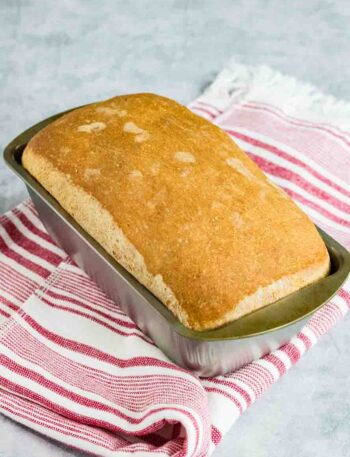 home made sandwich bread still sitting in the silver loaf pan on top of a red and white striped kitchen towel