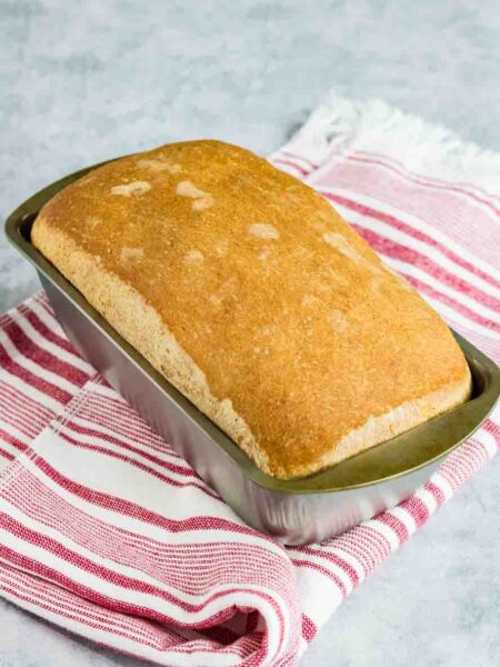 home made sandwich bread still sitting in the silver loaf pan on top of a red and white striped kitchen towel