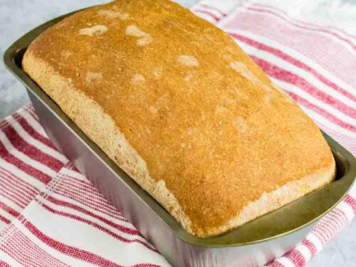 home made sandwich bread still sitting in the silver loaf pan on top of a red and white striped kitchen towel