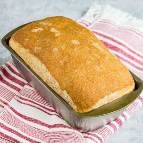 home made sandwich bread still sitting in the silver loaf pan on top of a red and white striped kitchen towel