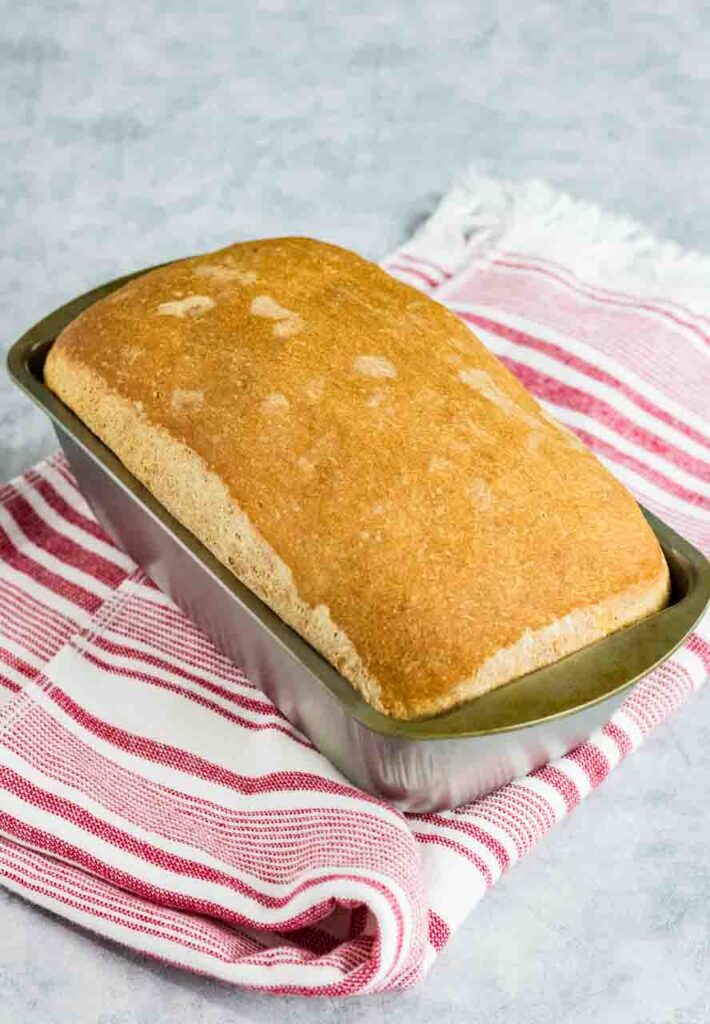 home made sandwich bread still sitting in the silver loaf pan on top of a red and white striped kitchen towel