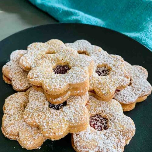 a black plate full of Linzer Tarts Cookies and a green towel in the background