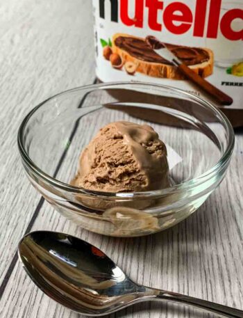 A scoop of Nutella Ice Cream sitting in a glass bowl with the Nutella Container behind it and a spoon in front of it