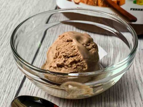 A scoop of Nutella Ice Cream sitting in a glass bowl with the Nutella Container behind it and a spoon in front of it