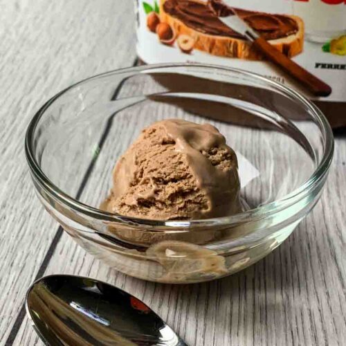 A scoop of Nutella Ice Cream sitting in a glass bowl with the Nutella Container behind it and a spoon in front of it