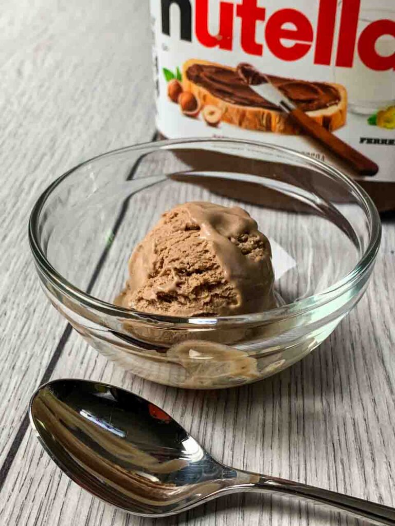 A scoop of Nutella Ice Cream sitting in a glass bowl with the Nutella Container behind it and a spoon in front of it