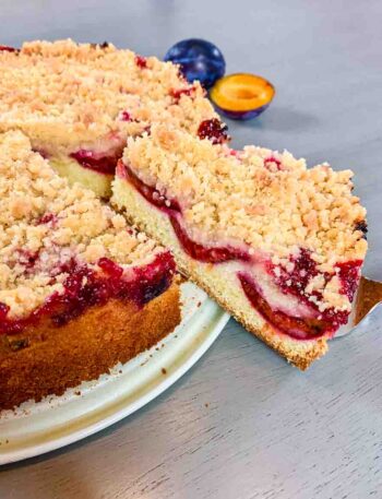 German plum cake as a whole on a cake platter with one piece sticking out on a cake shovel.