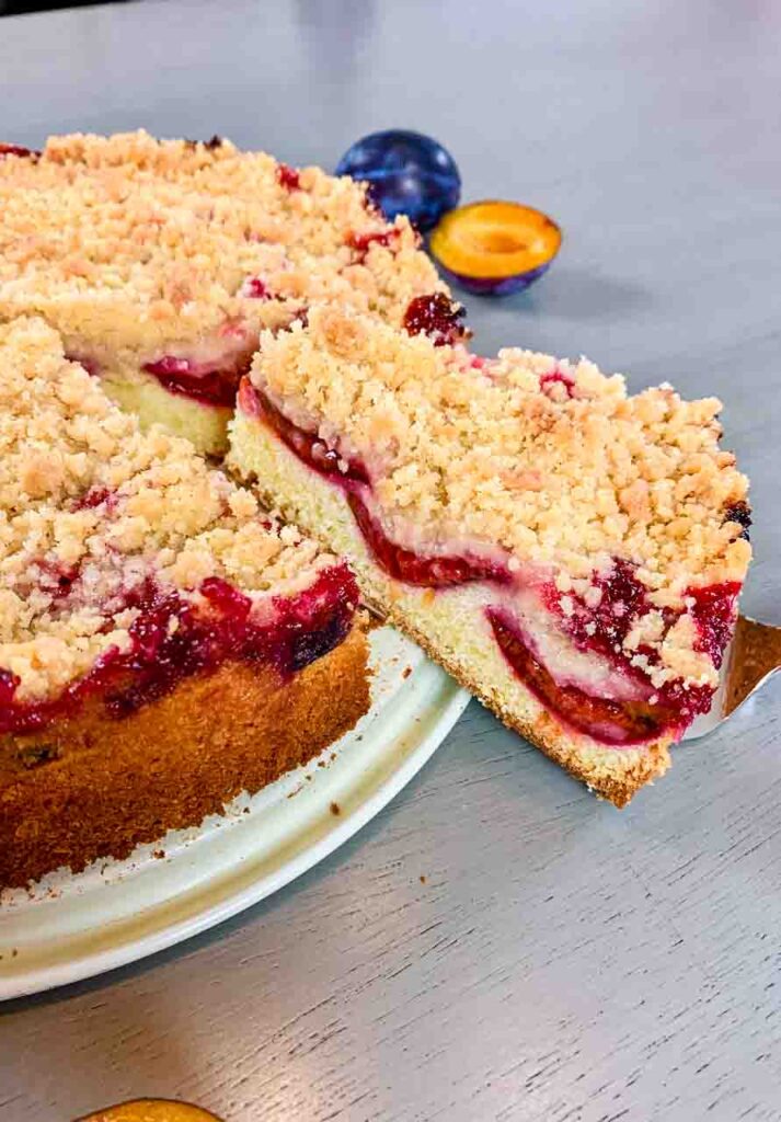 German plum cake as a whole on a cake platter with one piece sticking out on a cake shovel.