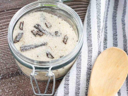 homemade vanilla sugar in a glass with a lid to seal it. Also visible are 3 vanilla beans and a wooden spoon on top of a dish towel