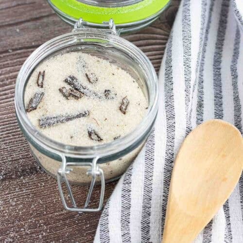 homemade vanilla sugar in a glass with a lid to seal it. Also visible are 3 vanilla beans and a wooden spoon on top of a dish towel
