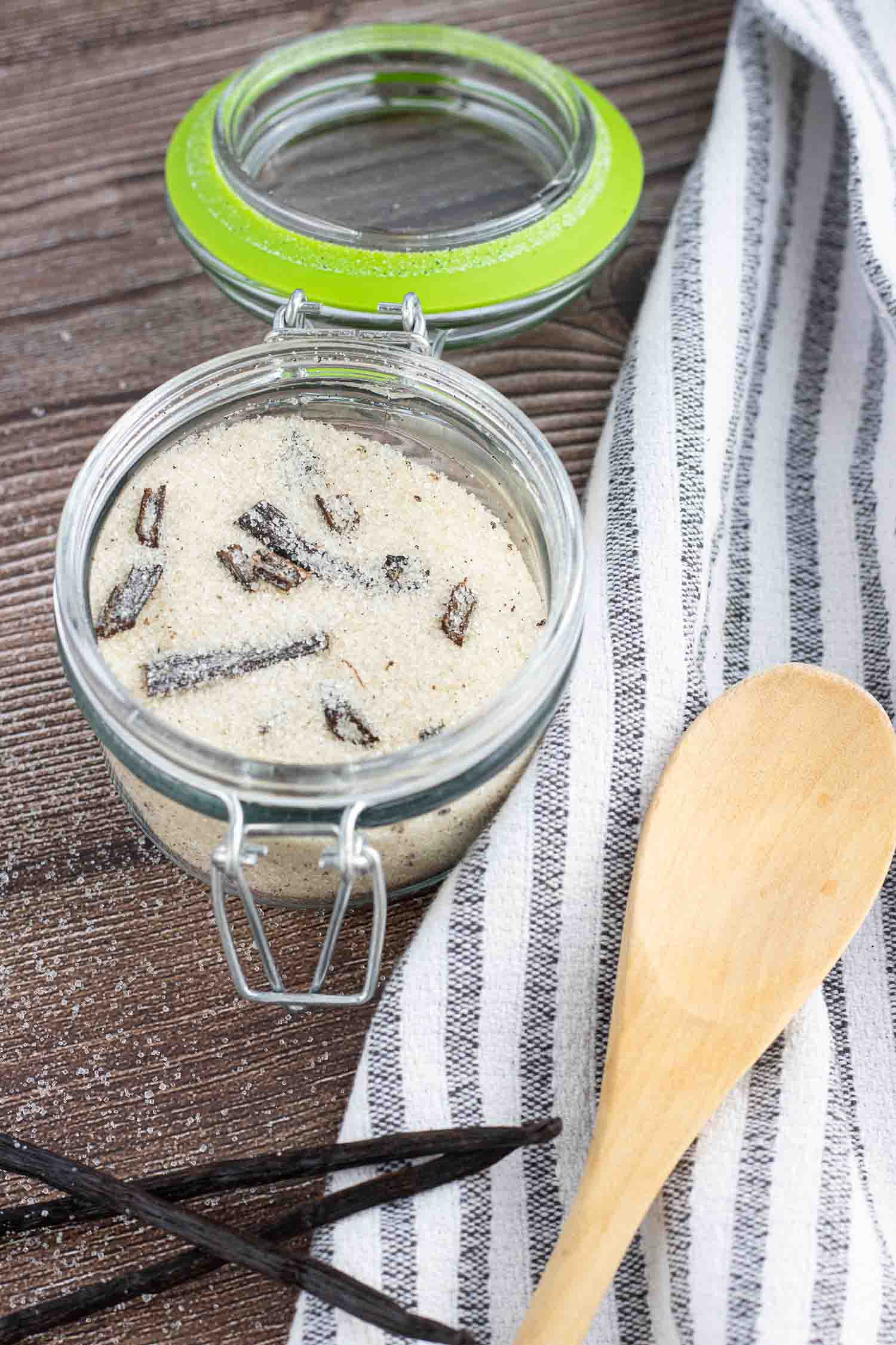 homemade vanilla sugar in a glass with a lid to seal it. Also visible are 3 vanilla beans and a wooden spoon on top of a dish towel