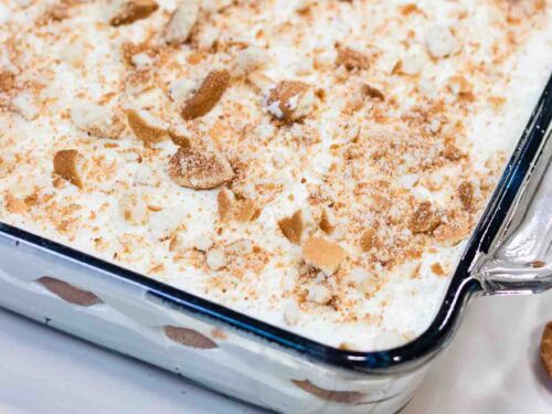 Banana Pudding on a square Casserole and crushed vanilla wafers on top. Banana slices and wafer are laying on the side of the casserole