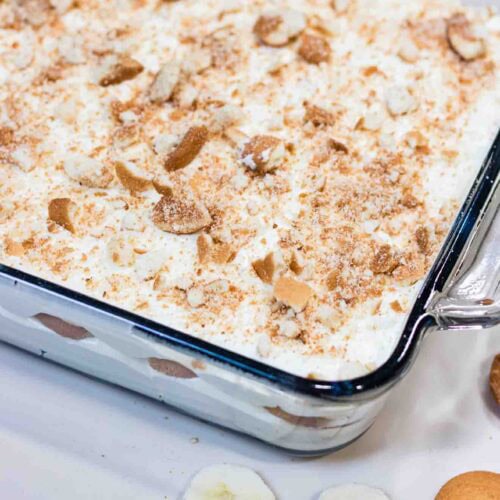 Banana Pudding on a square Casserole and crushed vanilla wafers on top. Banana slices and wafer are laying on the side of the casserole