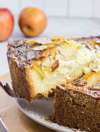Apple cake on a plate with on slice elevated on a cake spoon. It shows its moist inside and has almond slices on the top.