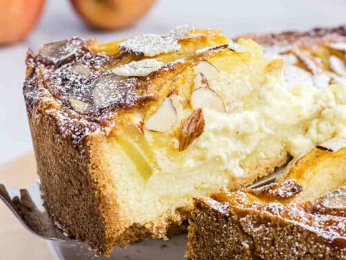 Apple cake on a plate with on slice elevated on a cake spoon. It shows its moist inside and has almond slices on the top.