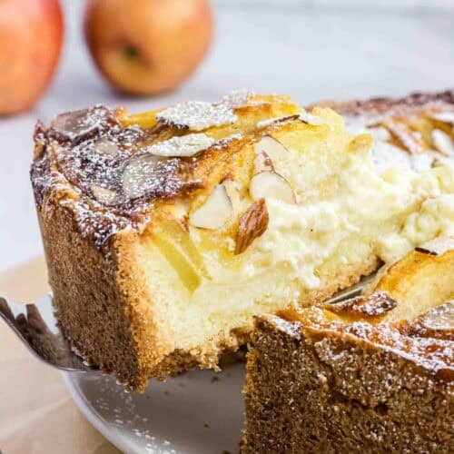 Apple cake on a plate with on slice elevated on a cake spoon. It shows its moist inside and has almond slices on the top.