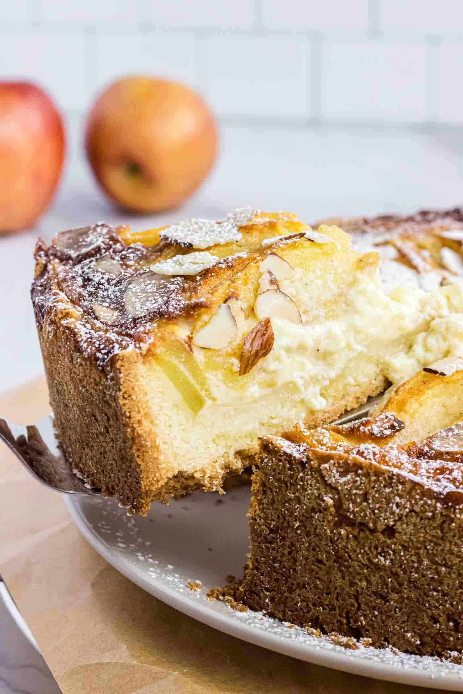 Apple cake on a plate with on slice elevated on a cake spoon. It shows its moist inside and has almond slices on the top.