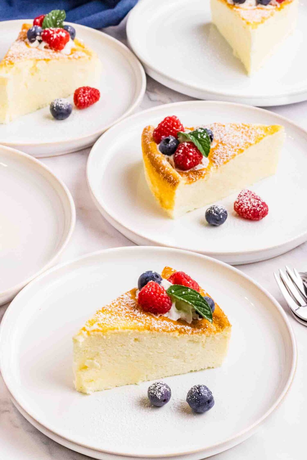 cake pieces on white plates spread out on the table. Each piece is decorated with 2 raspberries, blueberries, mint leaves and a swirl of whipping cream. Some raspberries and blueberries are also on the side of the cake on the plates.