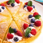 Yogurt cake cut into 8 equal pieces and decorated with powdered sugar, a swirl of whipping cream, 2 raspberries, 2 blueberries and a mint leaf on each slice. There is a blue kitchen towel on the left top and a blue small cake plate on the right top corner.