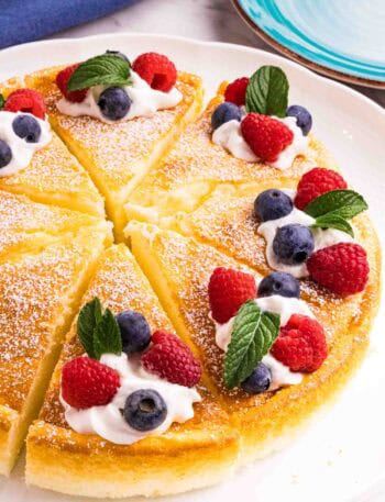 Yogurt cake cut into 8 equal pieces and decorated with powdered sugar, a swirl of whipping cream, 2 raspberries, 2 blueberries and a mint leaf on each slice. There is a blue kitchen towel on the left top and a blue small cake plate on the right top corner.