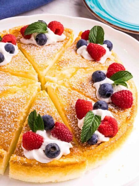 Yogurt cake cut into 8 equal pieces and decorated with powdered sugar, a swirl of whipping cream, 2 raspberries, 2 blueberries and a mint leaf on each slice. There is a blue kitchen towel on the left top and a blue small cake plate on the right top corner.