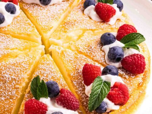 Yogurt cake cut into 8 equal pieces and decorated with powdered sugar, a swirl of whipping cream, 2 raspberries, 2 blueberries and a mint leaf on each slice. There is a blue kitchen towel on the left top and a blue small cake plate on the right top corner.