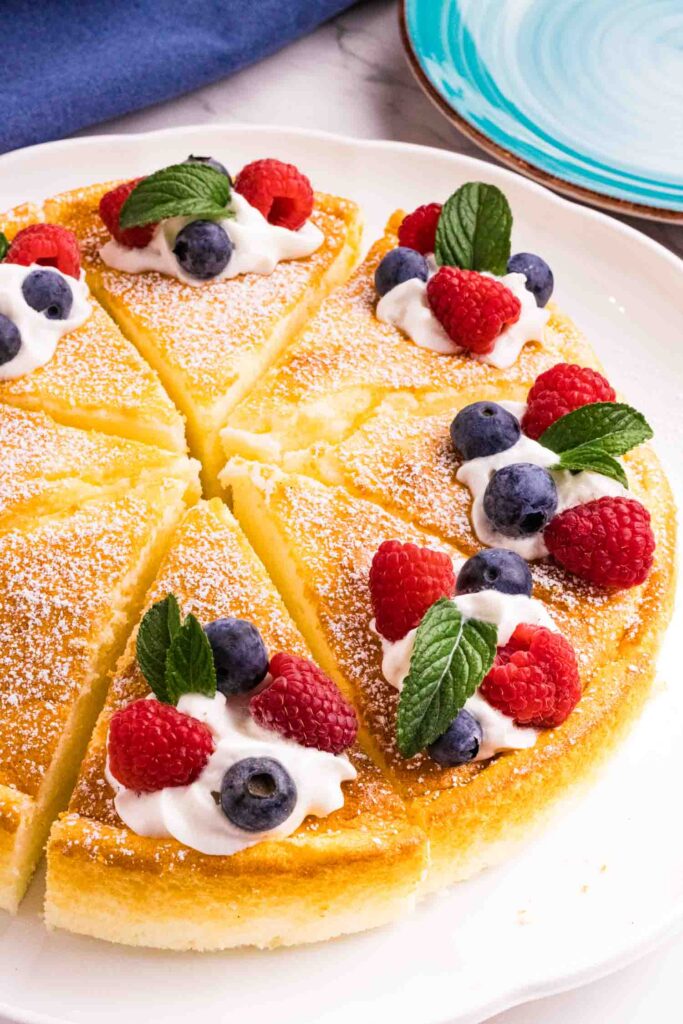 Yogurt cake cut into 8 equal pieces and decorated with powdered sugar, a swirl of whipping cream, 2 raspberries, 2 blueberries and a mint leaf on each slice. There is a blue kitchen towel on the left top and a blue small cake plate on the right top corner.