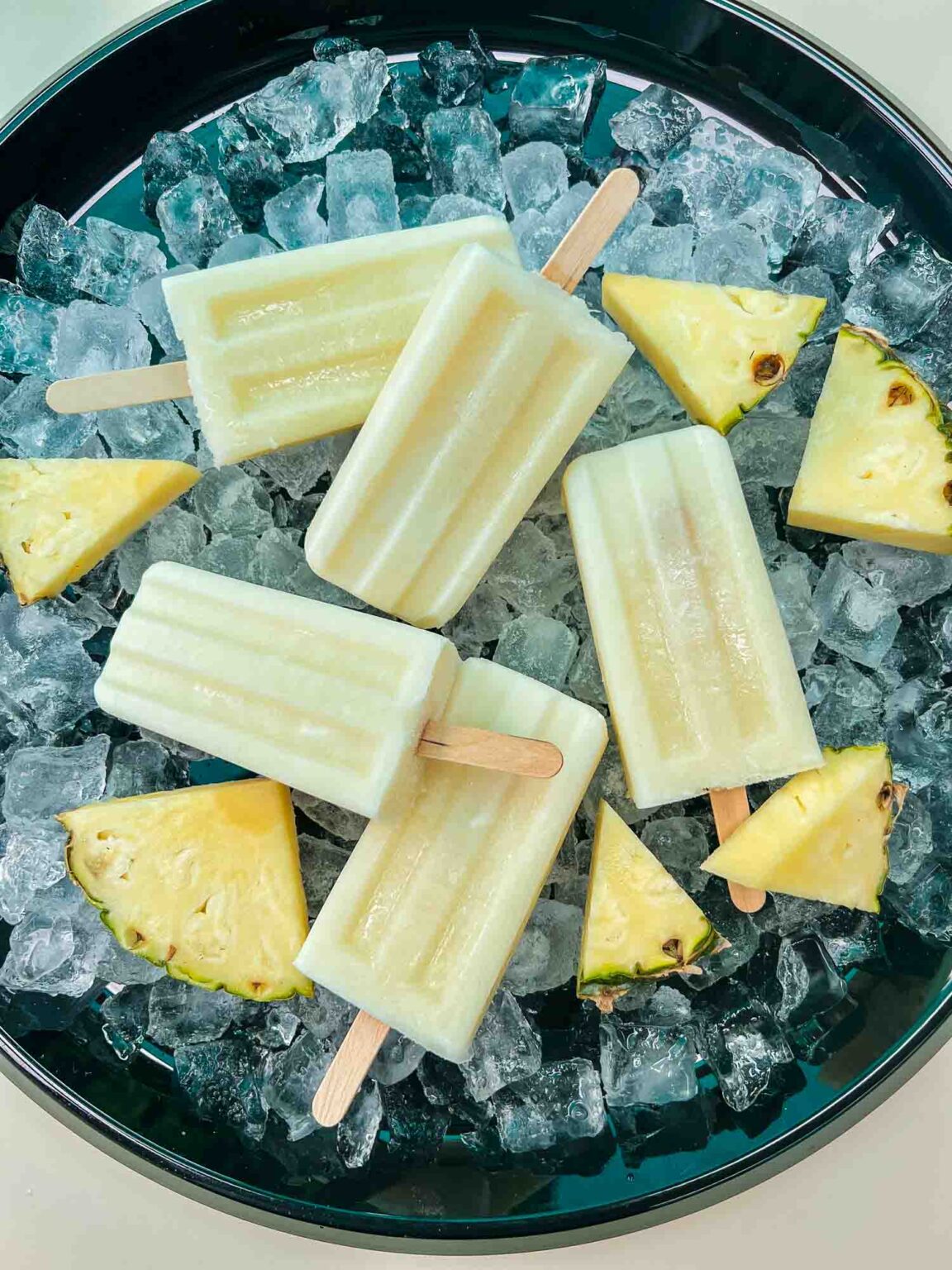 5 Pina Colada Popsicles on a bed of ice in a black tray surrounded by pineapple pieces