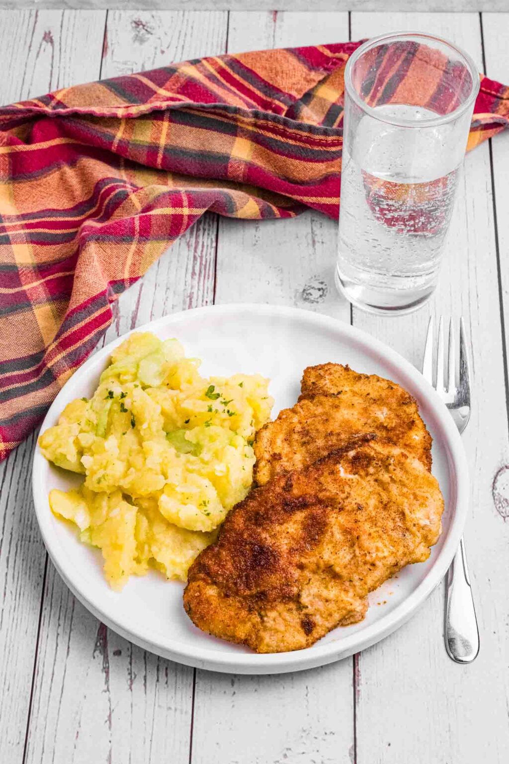 white rimmed plate with 2 pieces of Wiener schnitzel and potato salad on the side. beside it a fork, glass of water and a red plaid kitchen towel.