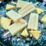 Pina Colada Popcicles laying on a bed of ice, surrounded by fresh pineapple pieces on a black round tray