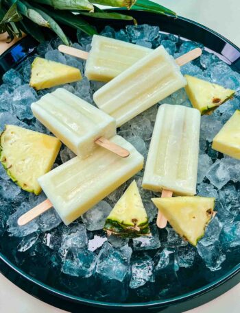 Pina Colada Popcicles laying on a bed of ice, surrounded by fresh pineapple pieces on a black round tray