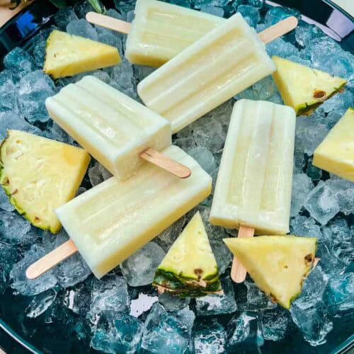 Pina Colada Popcicles laying on a bed of ice, surrounded by fresh pineapple pieces on a black round tray
