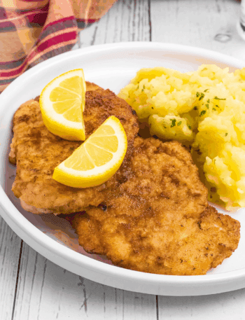 2 breaded Schnitzel pieces beside a pile of German potato salad. It is on a white rimmed plate and meat has two lemon slices on top.