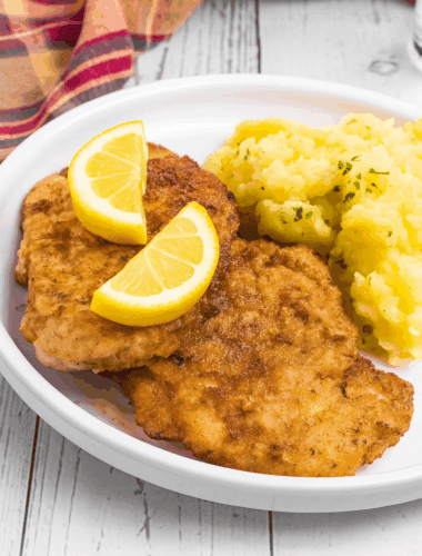 2 breaded Schnitzel pieces beside a pile of German potato salad. It is on a white rimmed plate and meat has two lemon slices on top.