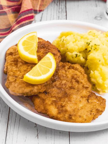2 breaded Schnitzel pieces beside a pile of German potato salad. It is on a white rimmed plate and meat has two lemon slices on top.