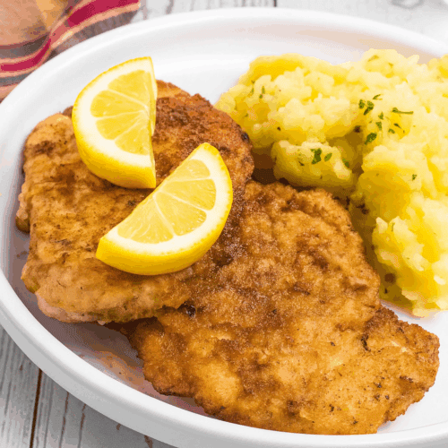 2 breaded Schnitzel pieces beside a pile of German potato salad. It is on a white rimmed plate and meat has two lemon slices on top.