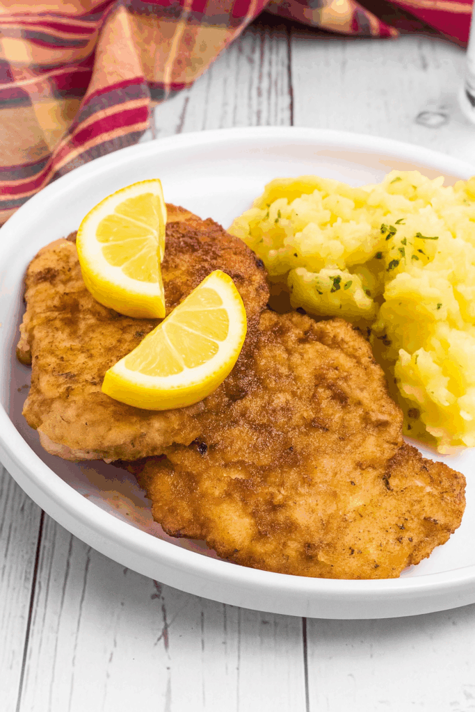 2 breaded Schnitzel pieces beside a pile of German potato salad. It is on a white rimmed plate and meat has two lemon slices on top.