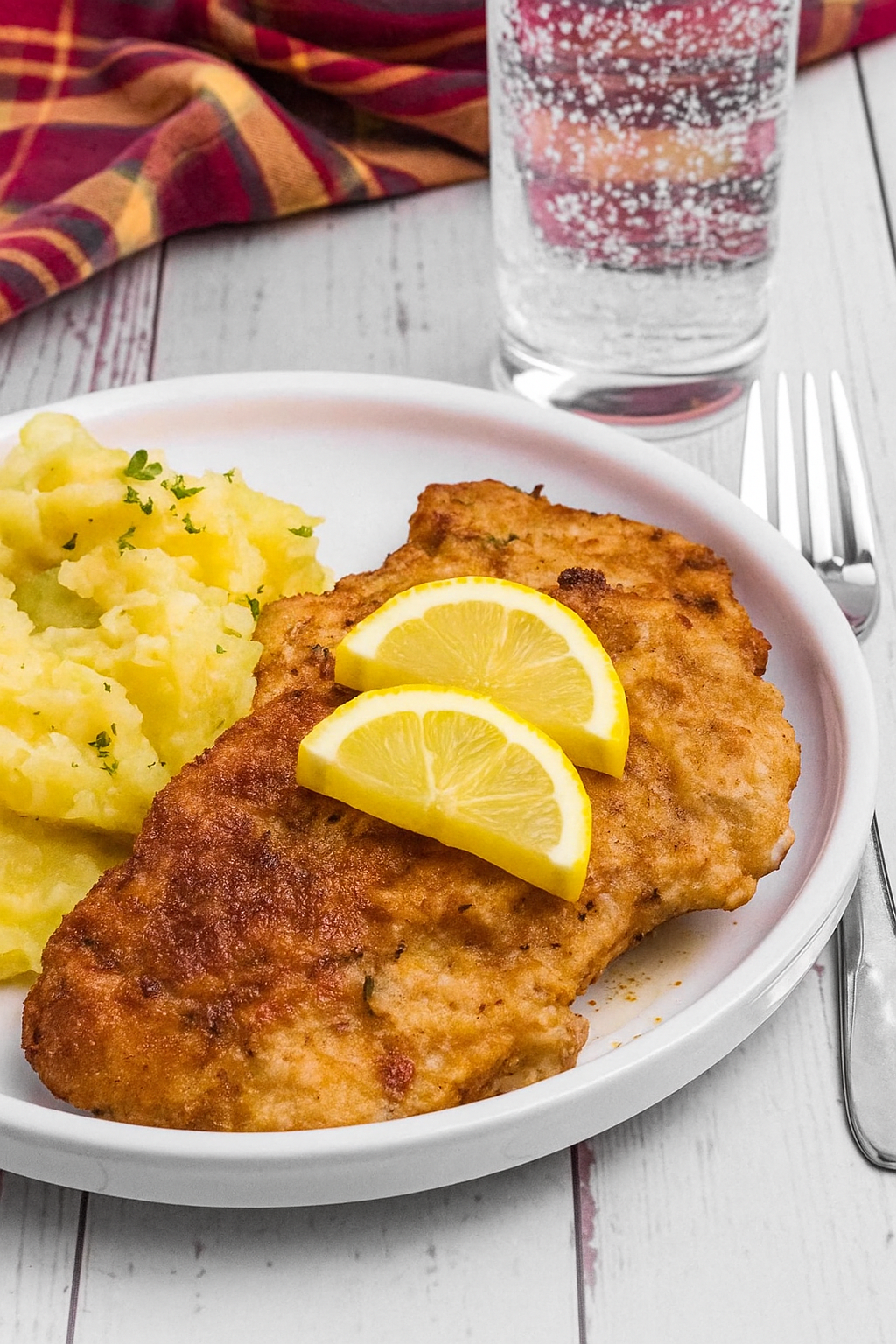 White plate with 2 breaded pieces of Wiener schnitzel and a big spoon of potato salad. Beside it is a fork, glass of water and a red plait kitchen towel. It sits on a white wooden table.