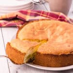 whole golden brown rhubarb cake with on slice slightly elevated on a cake shovel. Cups, plates and forks are in the background.